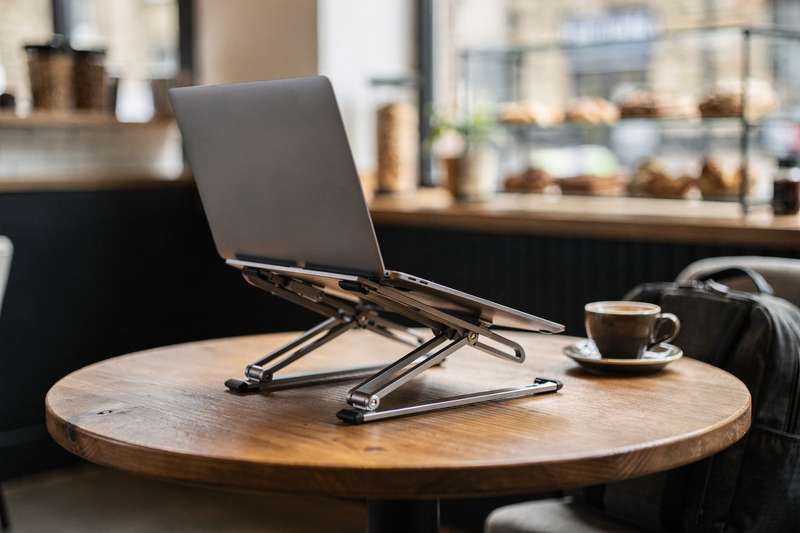 Compact foldable laptop stand displayed next to a work bag and coffee cup in a café setting.