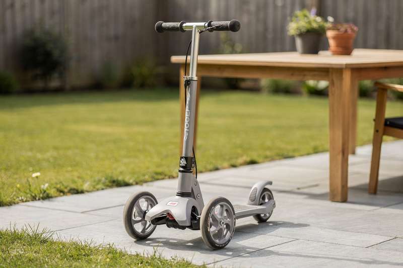 Child performing a trick on a three-wheeled scooter in an outdoor park setting.