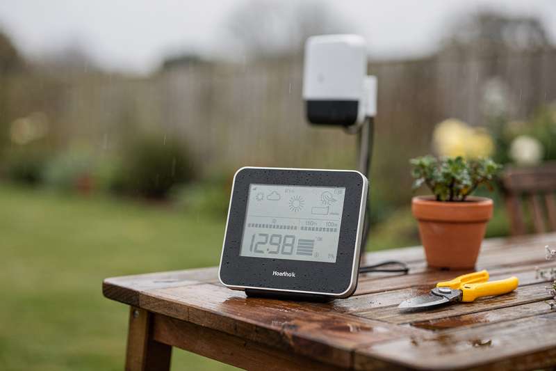 Digital and analog weather station models displayed on a garden fence with rain gauge and thermometer attachments.
